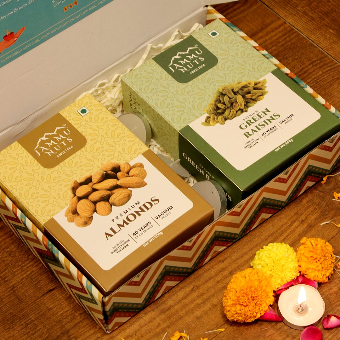 Two boxes of Ammu Nuts, one with almonds and the other with green raisins, on a wooden surface with decorative flowers and a candle.