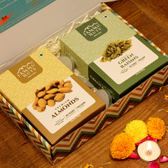 Two boxes of Ammu Nuts, one with almonds and the other with green raisins, on a wooden surface with decorative flowers and a candle.